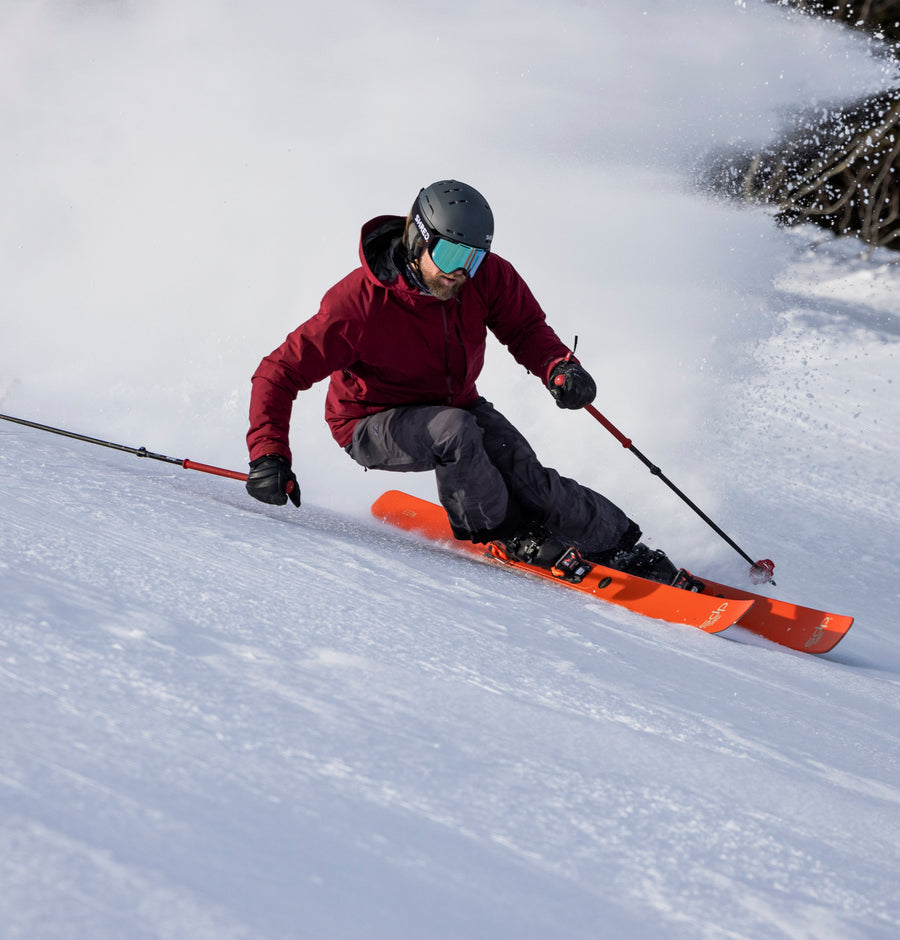 Person skiing down a snowy slope with orange skis and red jacket.